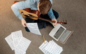 موسيقي يراجع عملية التدوين الموسيقي باستخدام حاسوبه وبعض الاوراق لنوتات موسيقية مختلفة وهو يحمل جيتاره